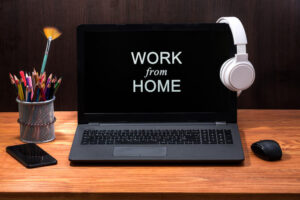 An open black laptop sits on a wooden desk with a pair of white headphones hanging from the open screen. There is a black computer mouse to the right. On the left is a cup full of pencil crayons and a fan paint brush, and a mobile phone next to it.