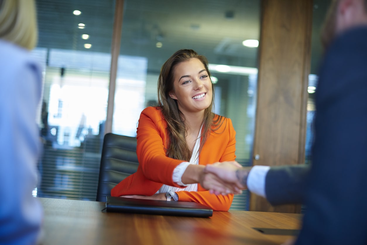 A young woman sits at a table across from a job interviewer and gives them a handshake.