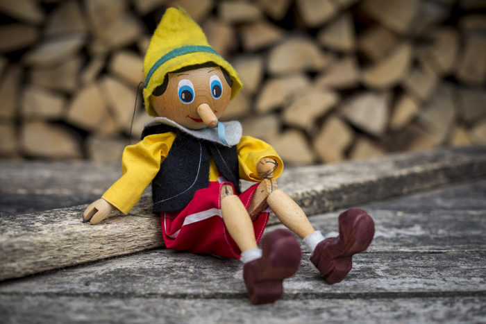 A wooden Pinocchio figure with his signature long nose sits on a wooden background.