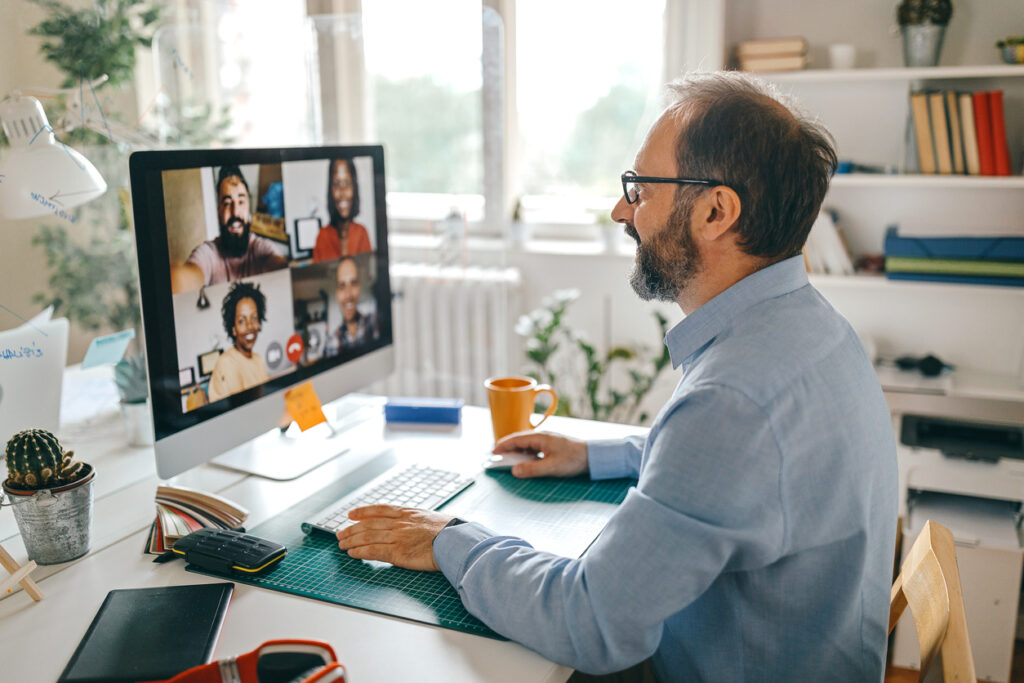A man in glasses works from his home office while video chatting with his coworkers through his computer.