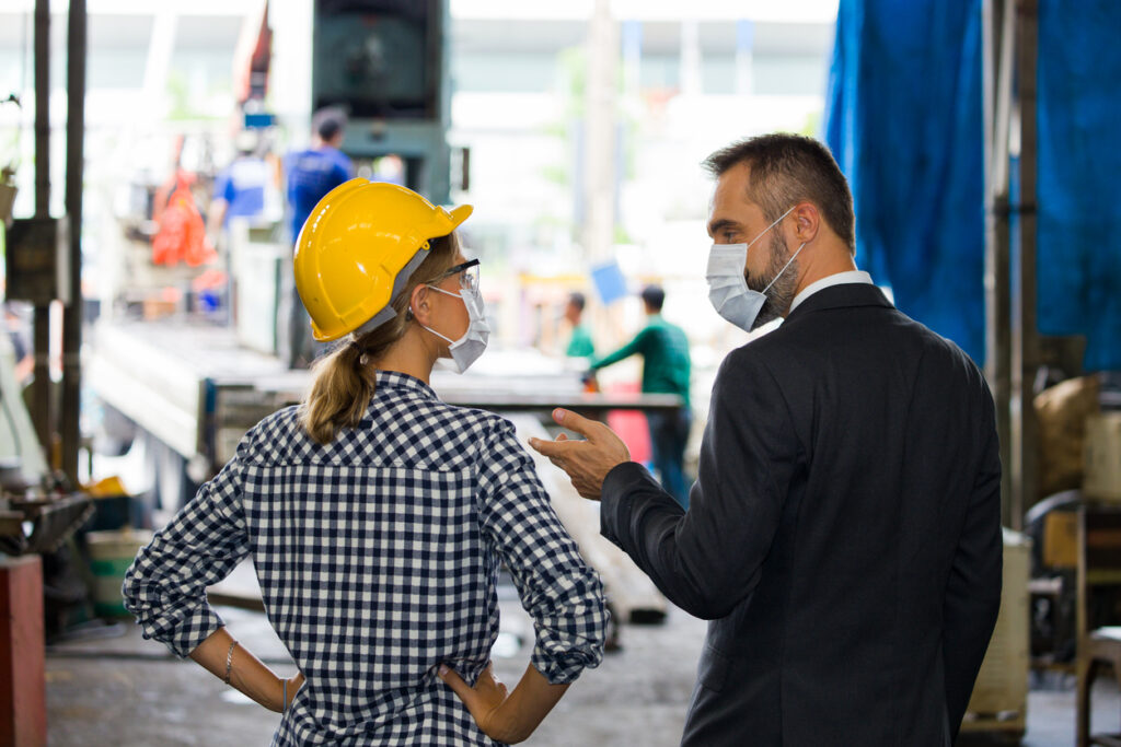 A woman engineer in a yellow hardhat and a businessman are both wearing medical masks while discussing a manufacturing project.